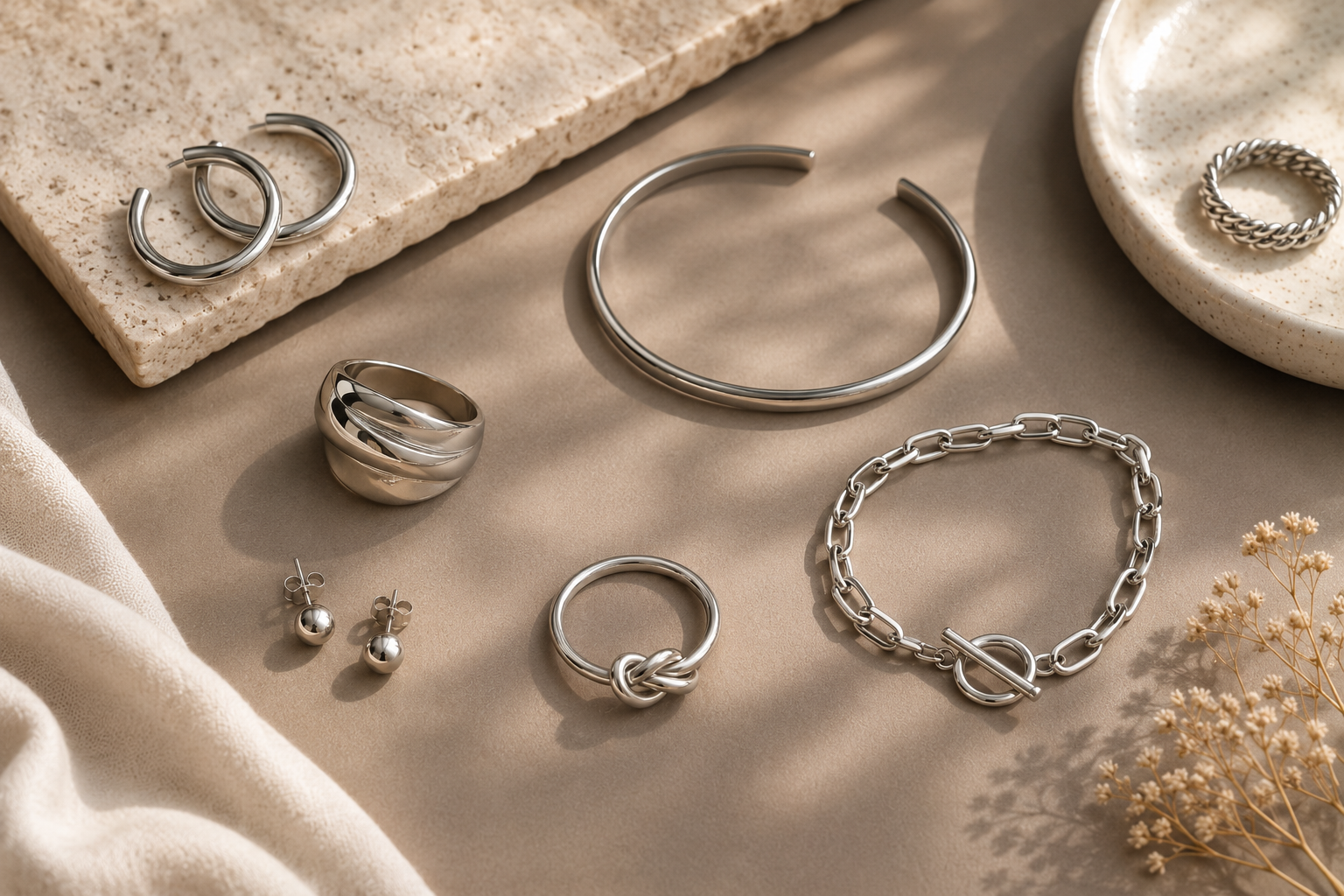 Composição elegante de joias prateadas femininas sobre fundo taupe sofisticado, com anéis, brincos, pulseira e bracelete iluminados por luz natural suave.